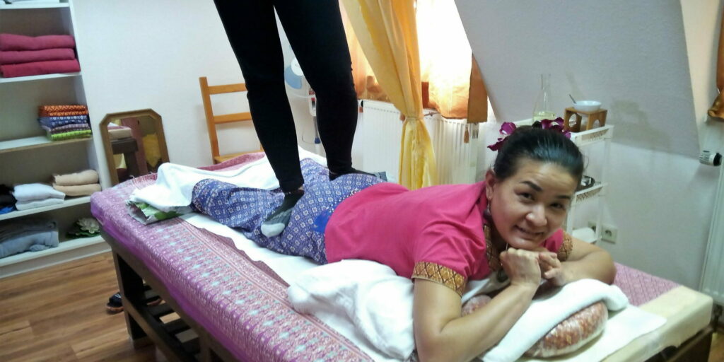 Eine Frau mit rosa T-Shirt liegt auf dem Bauch auf einer Massageliege und lächelt in die Kamera.