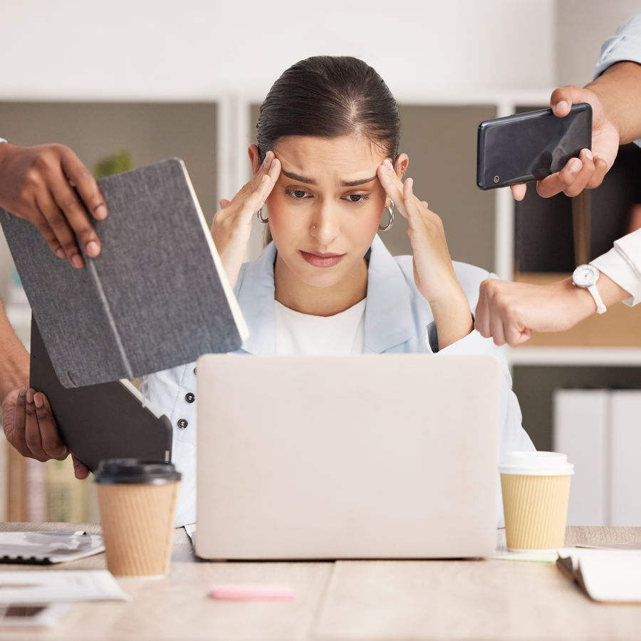 Eine junge Geschäftsfrau sitzt an einem Schreibtisch hinter einem Laptop und hält die Hände an die Schläfen, da sie unter Stress steht.