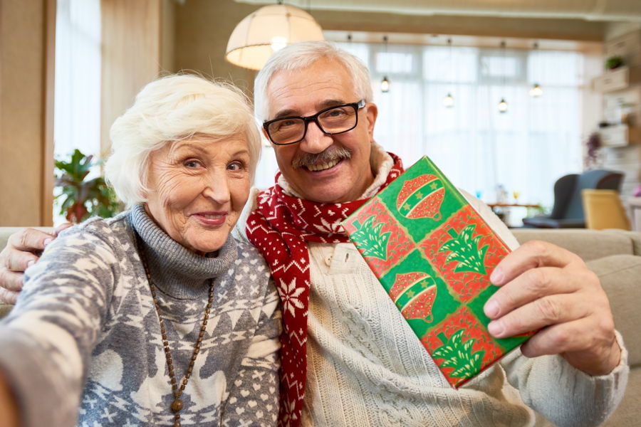 Ein fröhliches Seniorenpaar hält einen Massagegutschein in die Kamerq, den es als Weihnachtsgeschenk erhalten hat.