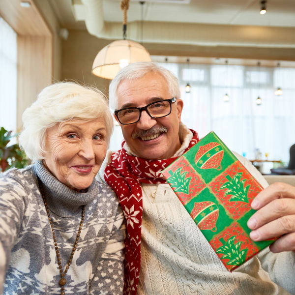 Ein fröhliches Seniorenpaar hält einen Massagegutschein in die Kamerq, den es als Weihnachtsgeschenk erhalten hat.